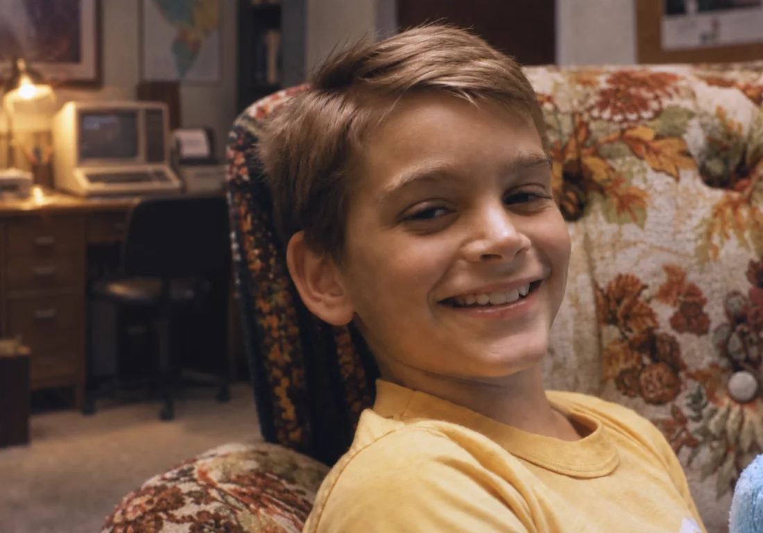James at six, grinning in a floral armchair with a TRS-80 computer on the desk behind him